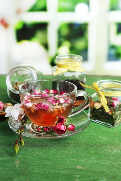 Sortimento de ervas, chá em frascos de vidro e bebida quente em xícara na mesa de madeira, no fundo brilhante — Fotografia de Stock