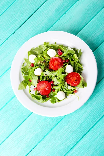 Grüner Salat mit Rucola, Tomaten, Käse-Mozzarella-Bällchen und Sesam auf Teller, auf farbigem Holzhintergrund — Stockfoto