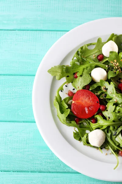 Grüner Salat mit Rucola, Tomaten, Käse-Mozzarella-Bällchen und Sesam auf Teller, auf farbigem Holzhintergrund — Stockfoto