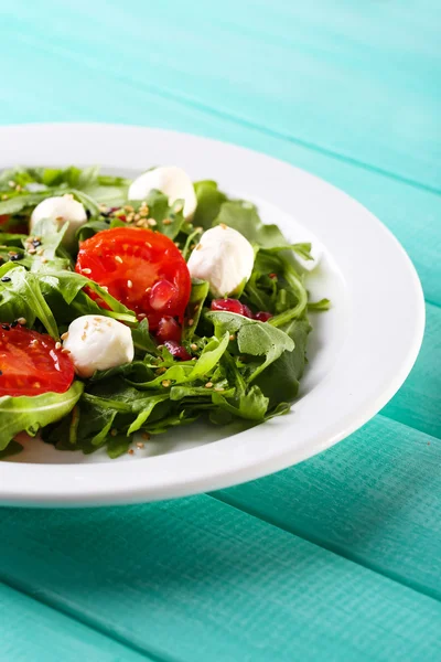 Groene salade gemaakt met rucola, tomaten, kaas mozzarella ballen en sesam op plaat, op een houten achtergrond kleur — Stockfoto