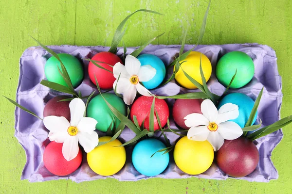 Barevné velikonoční vajíčka s trávou a květinami v zásobníku na dřevěné pozadí — Stock fotografie