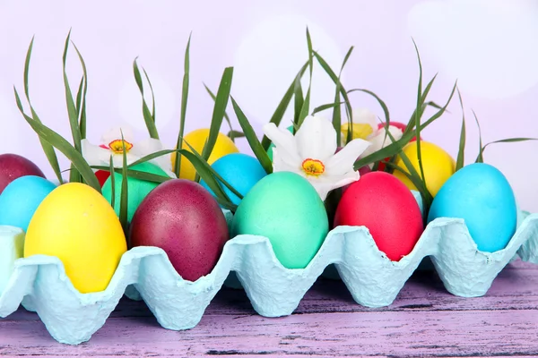 Barevné velikonoční vajíčka s trávou a květinami v zásobníku na stole na světlé pozadí — Stock fotografie