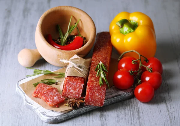 Tasty salami sausage, vegetables and spices on paper, on wooden background — Stock Photo, Image