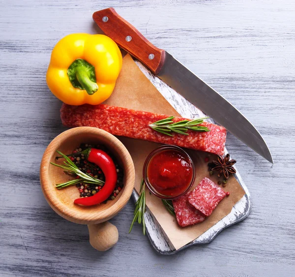 Salsicha de salame saborosa, legumes e especiarias no papel, no fundo de madeira — Fotografia de Stock