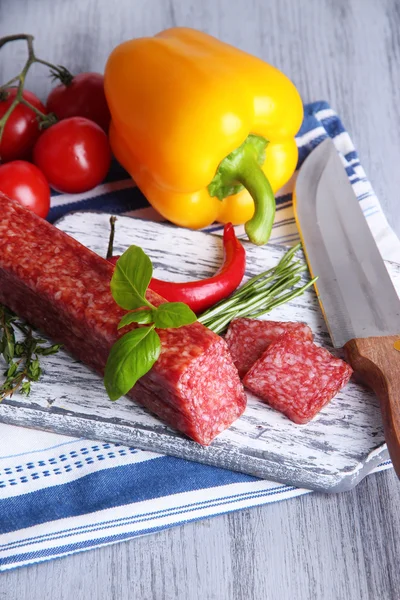 Leckere Salami-Wurst, Gemüse, Messer und Gewürze auf Holzgrund — Stockfoto