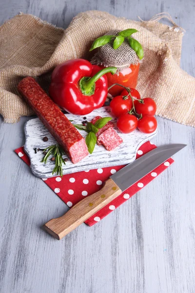 Tasty salami sausage, vegetables, knife and spices on wooden background — Stock Photo, Image