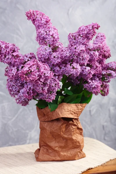 Beautiful lilac flowers in paper bag on wooden table near wall — Stock Photo, Image