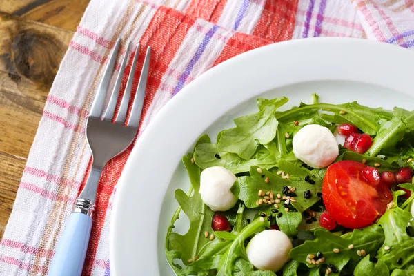 Grüner Salat mit Rucola, Tomaten, Käse-Mozzarella-Bällchen und Sesam auf Teller, auf Holzgrund — Stockfoto