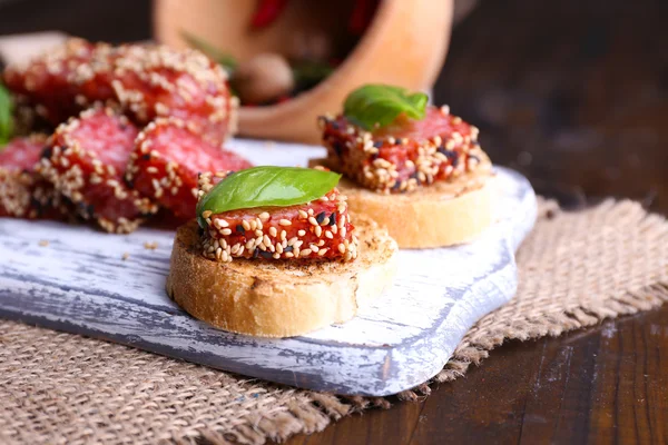 Composizione con coltello, gustosi panini con salsiccia di salame, foglia di basilico sul tagliere, su fondo di legno — Foto Stock