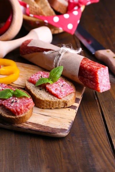 Composição com faca, sanduíches saborosos com salsicha de salame, folha de manjericão na tábua de corte, no fundo de madeira — Fotografia de Stock