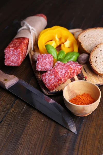 Composizione con coltello, saporita salsiccia di salame, pane e pepe a fette sul tagliere, su fondo di legno — Foto Stock