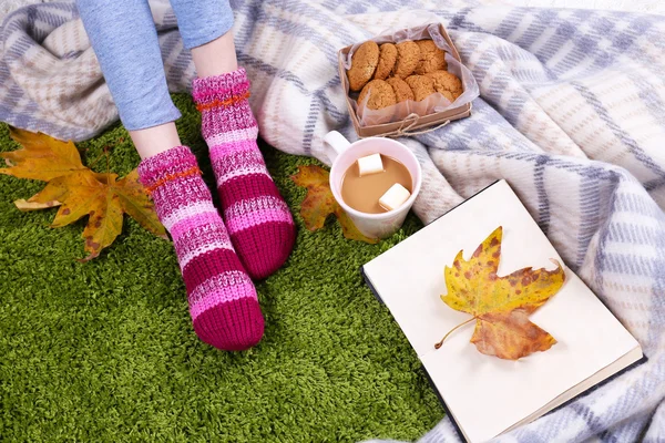 Composizione con plaid caldo, libro, tazza di bevanda calda e gambe femminili, su sfondo tappeto di colore — Foto Stock