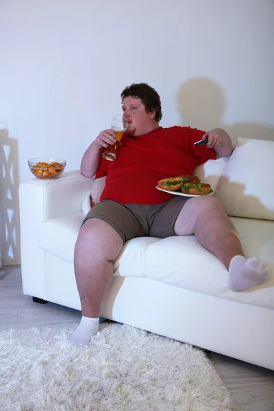 Hombre perezoso con sobrepeso sentado con un vaso de cerveza en el sofá y viendo la televisión —  Fotos de Stock