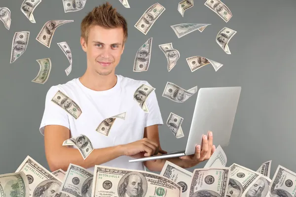 Mladý muž pracující na notebooku s deštěm peníze na šedém pozadí — Stock fotografie