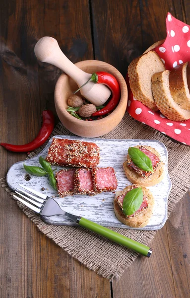 Sammansättning med kniv, läckra smörgåsar med salami korv, basilika blad på skärbräda, på trä bakgrund — Stockfoto