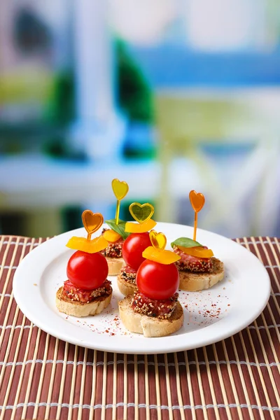 Välsmakande kanapéer med salami, tomat, paprika och basilika blad, på plattan, träbord, på ljus bakgrund — Stockfoto