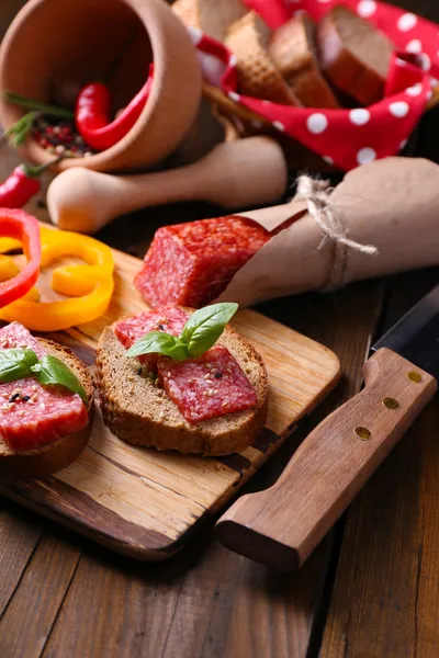 Samenstelling met mes, smakelijke sandwiches met salami worst, basilicum blad op snijplank, op houten achtergrond — Stockfoto