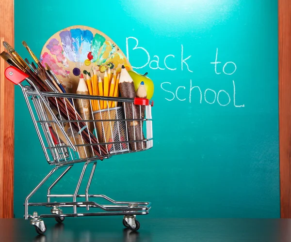 School supplies in supermarket cart on blackboard background — Stock Photo, Image