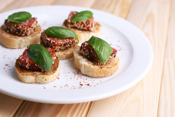 Gustose tartine con salumi e foglie di basilico, su piatto, su fondo di legno — Foto Stock