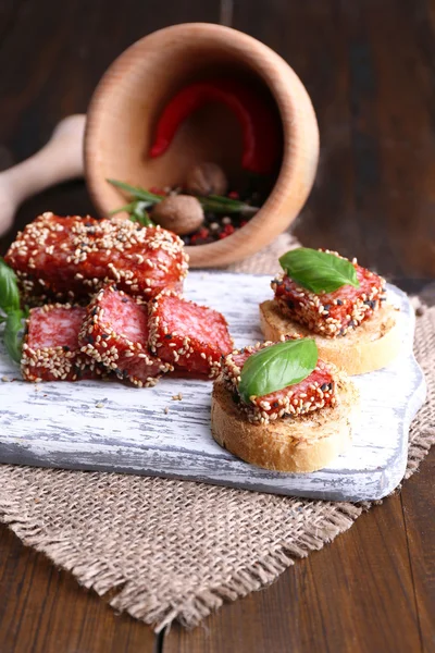 Sammansättning med kniv, läckra smörgåsar med salami korv, basilika blad på skärbräda, på trä bakgrund — Stockfoto