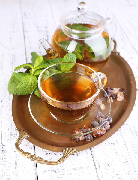 Состав свежего мятного чая в стеклянной чашке и чайник на подносе, на деревянном фоне
