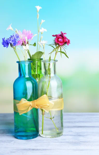 Fiori di campo lucenti in bottiglie su tavolo di legno, su sfondo chiaro — Foto Stock