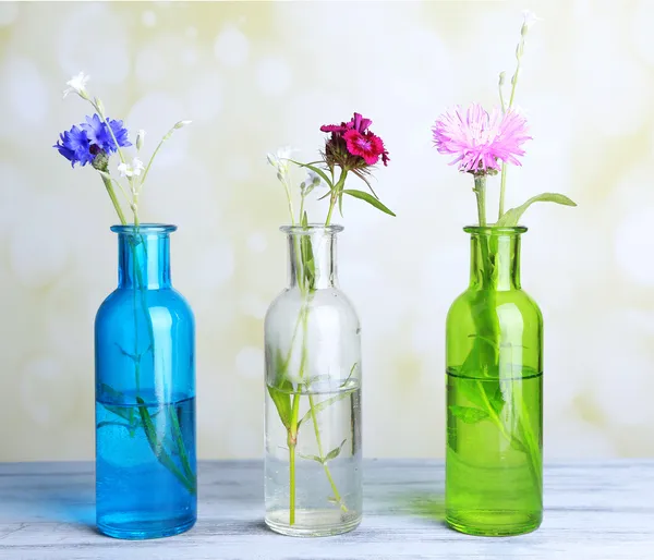 Heldere wilde bloemen in flessen op houten tafel, op lichte achtergrond — Stockfoto