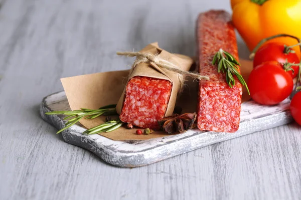 Tasty salami sausage, vegetables and spices on paper on wooden background — Stock Photo, Image