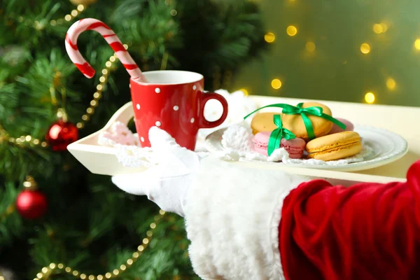 Santa sosteniendo taza y plato con galletas en la mano, sobre fondo brillante —  Fotos de Stock