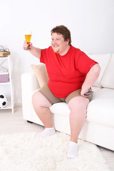 Hombre perezoso con sobrepeso sentado con un vaso de cerveza en el sofá y viendo la televisión —  Fotos de Stock