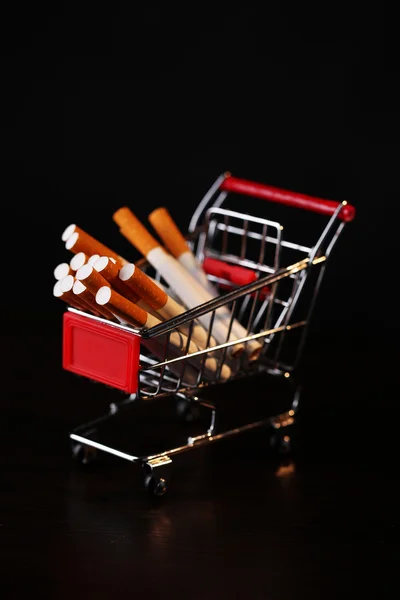 Cigarettes in shopping cart on wooden table on dark background — Stock Photo, Image