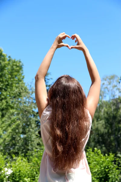 Молодая девушка держит руки в форме сердца, обрамляясь на фоне голубого неба — стоковое фото
