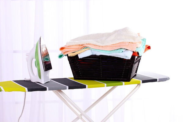 Basket with laundry and ironing board on light home interior background
