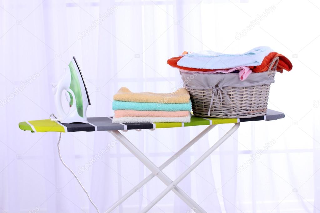 Basket with laundry and ironing board on light home interior background
