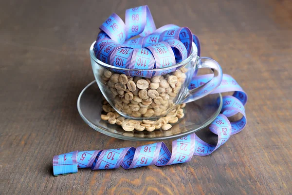 Raw green coffee beans in glass cup and measuring tape, on color wooden background. Concept of weight loss