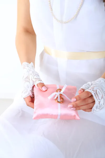 Nevěsta v bílých šatech a rukavice drží dekorativní polštář s snubních prstenů, close-up, na světlé pozadí — Stock fotografie