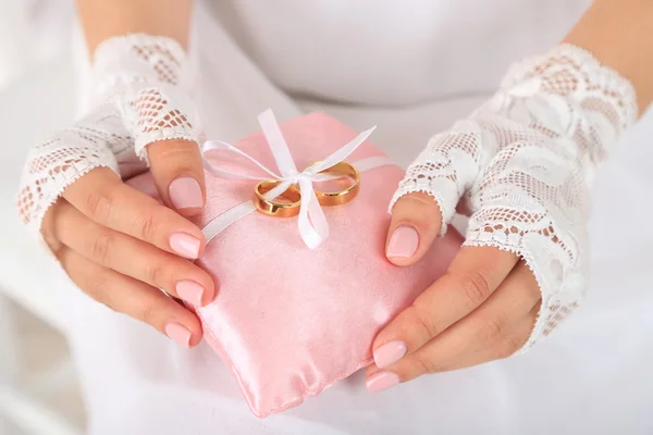 Noiva em vestido branco e luvas segurando travesseiro decorativo com anéis de casamento, close-up, no fundo claro — Fotografia de Stock