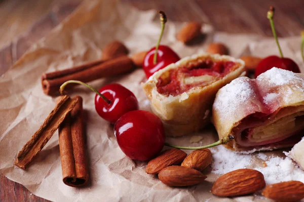 Tasty homemade strudel and fresh sweet cherry on paper napkin, on wooden table background — Stock Photo, Image