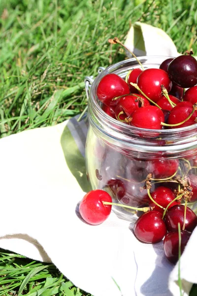 Cerises mûres douces dans un bocal en verre, sur une serviette, sur un fond d'herbe verte — Photo