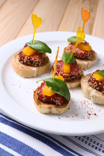 Tasty canapes with salami, pepper  and basil leaves, on plate, on wooden background — Stock Photo, Image