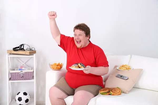 Hombre perezoso con sobrepeso sentado con comida rápida en el sofá y viendo la televisión —  Fotos de Stock