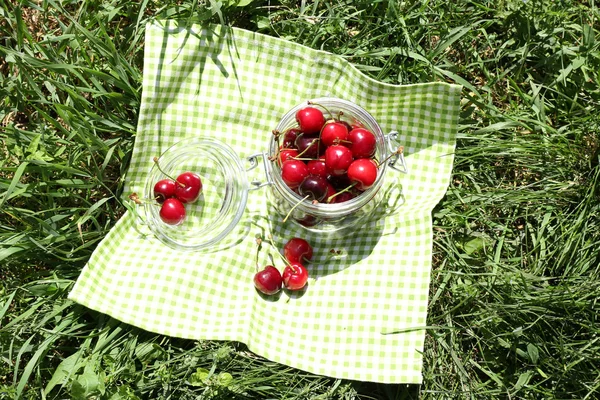 Сладкие спелые вишни в стеклянной банке, на салфетке, на зеленом фоне травы — стоковое фото