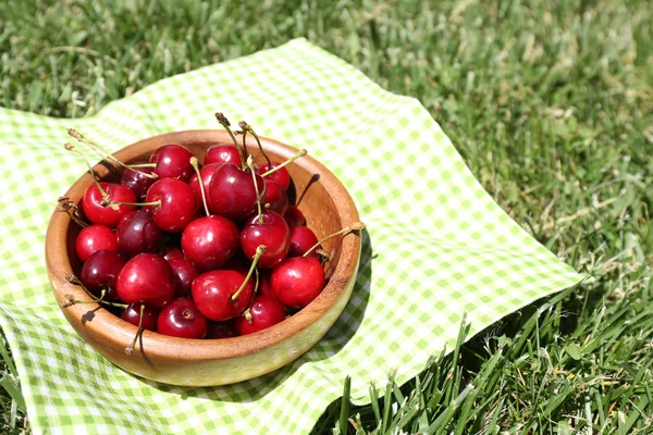 Sweet ripe cherries in wooden bowl, on napkin, on green grass background — Stock Photo, Image