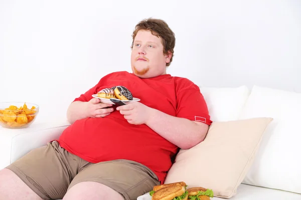 Preguiçoso excesso de peso masculino sentado com fast food no sofá e assistindo televisão — Fotografia de Stock