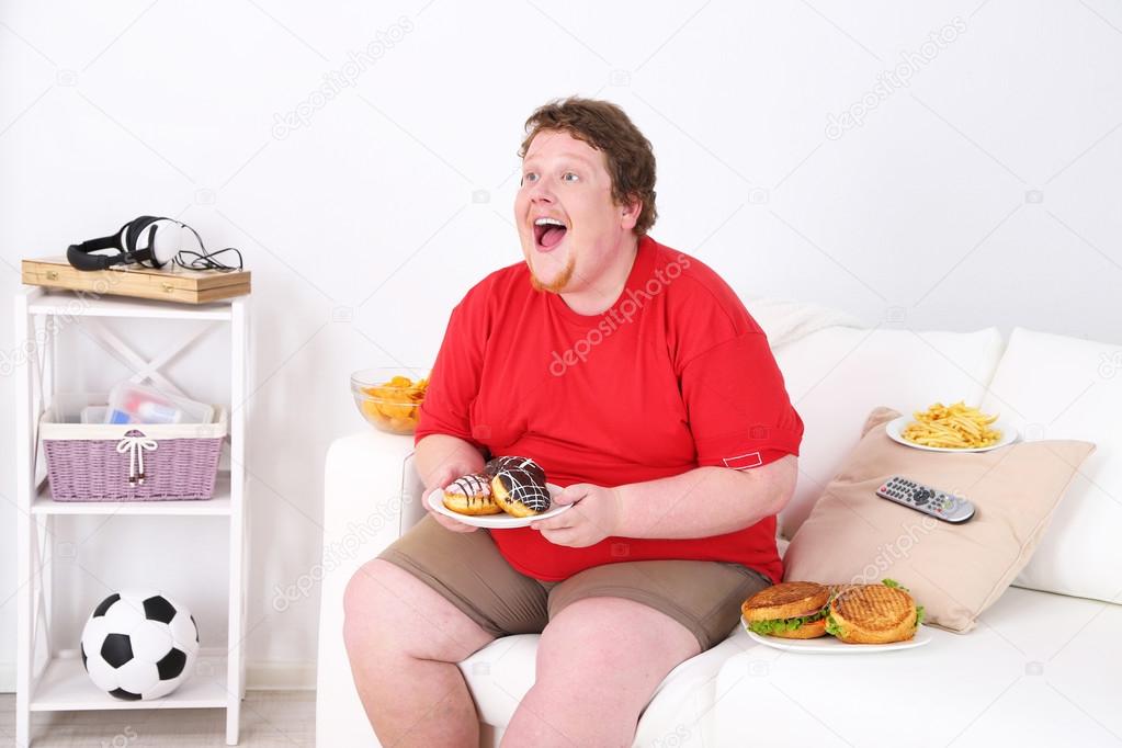 Lazy overweight male sitting with fast food on couch and watching television