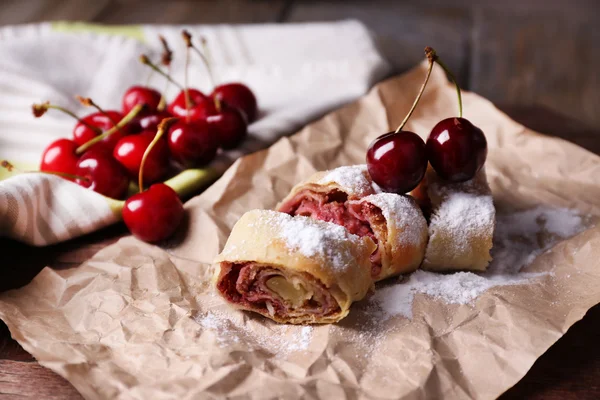 Leckerer hausgemachter Strudel und frische Süßkirsche auf Papierserviette, auf Holztischhintergrund — Stockfoto