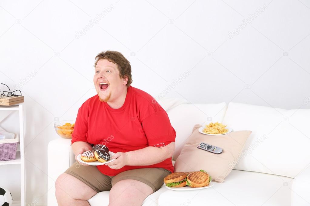 Lazy overweight male sitting with fast food on couch and watching television