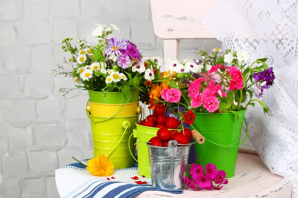 Bouquet of colorful flowers and fresh cherries in decorative buckets, on chair, on light wall background — Stock Photo, Image