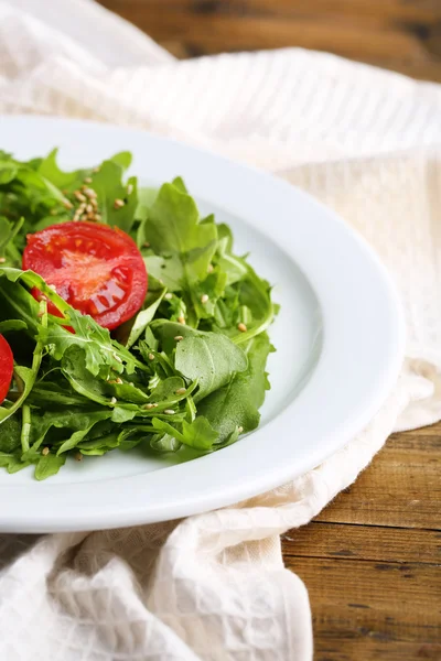 Зеленый салат из рукколы, помидоров и кунжута на тарелке, на деревянном фоне — стоковое фото