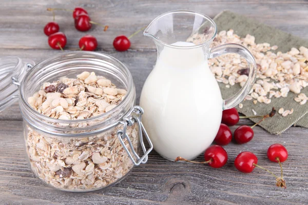 Homemade granola in glass jar, fresh cherries and jug with milk on color wooden background — Stock Photo, Image